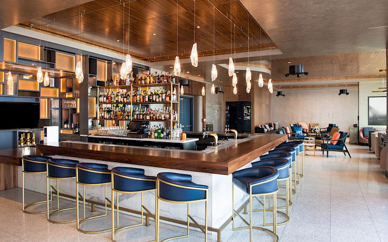 The image shows a modern bar with a sleek counter, blue chairs, pendant lights, and a fully stocked shelf of drinks behind the bar, ending the sentence.