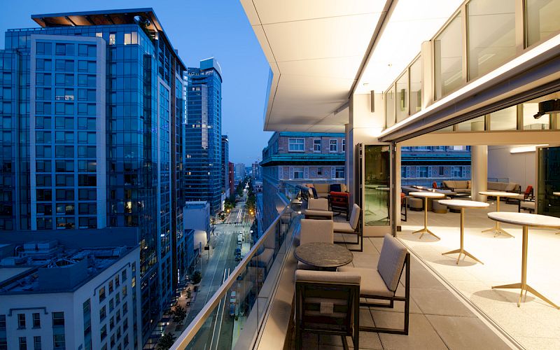The image shows a cityscape with high-rise buildings at dusk. A modern rooftop patio with chairs, tables, and glass railings overlooks the street.