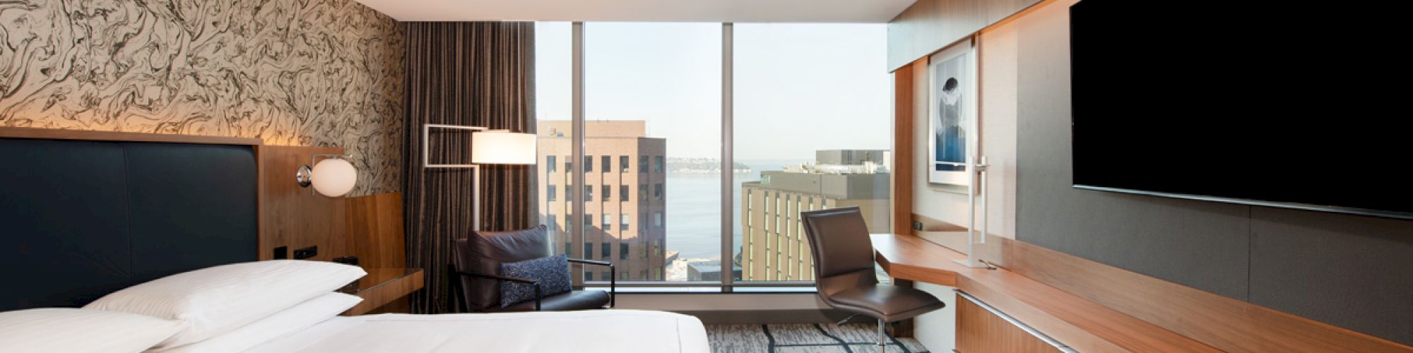 A modern hotel room with a large bed, desk, chair, big-screen TV, and a view of buildings and water through a floor-to-ceiling window.